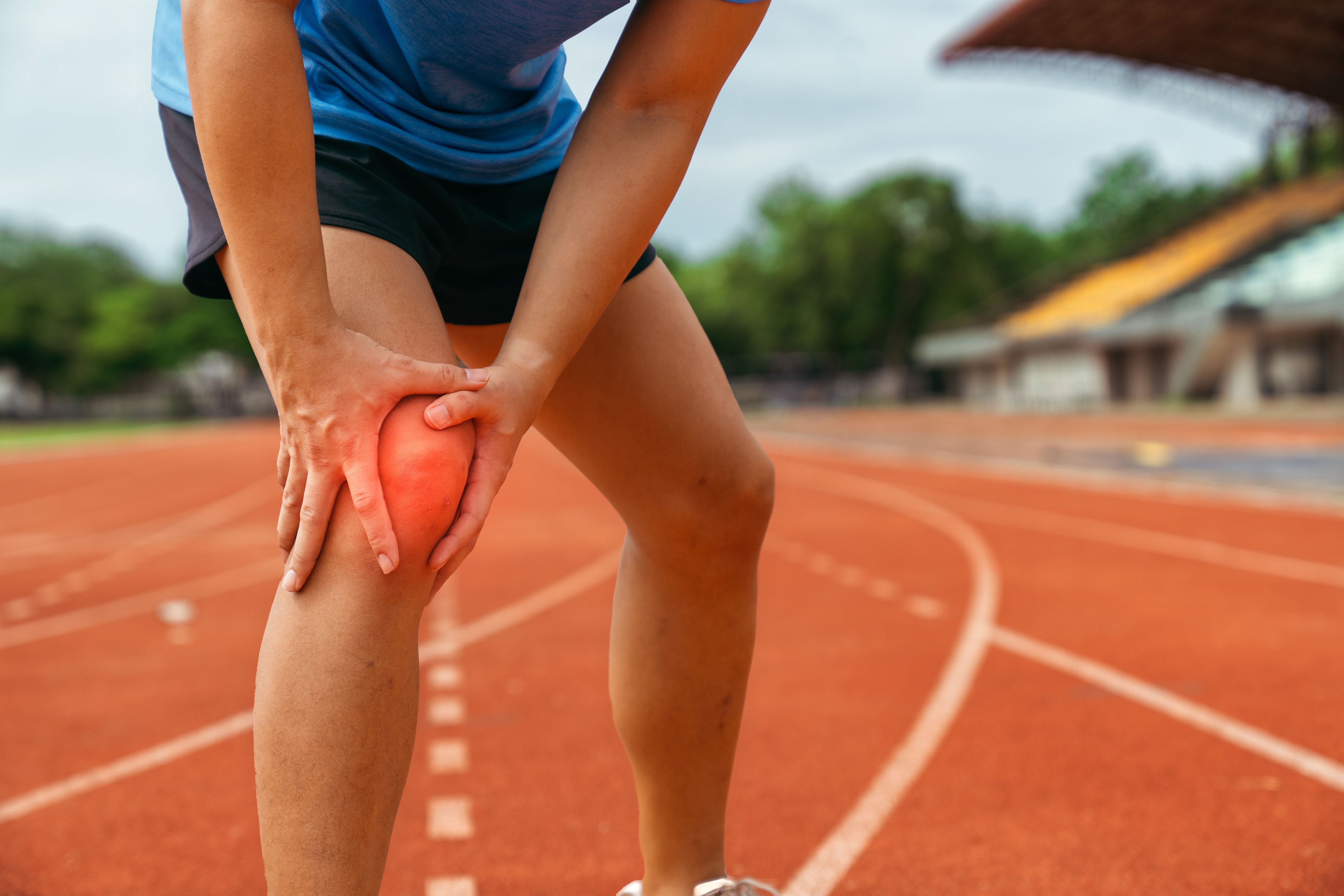 Runner clutching their knee in pain on an outdoor track, with a red highlight indicating injury or inflammation.