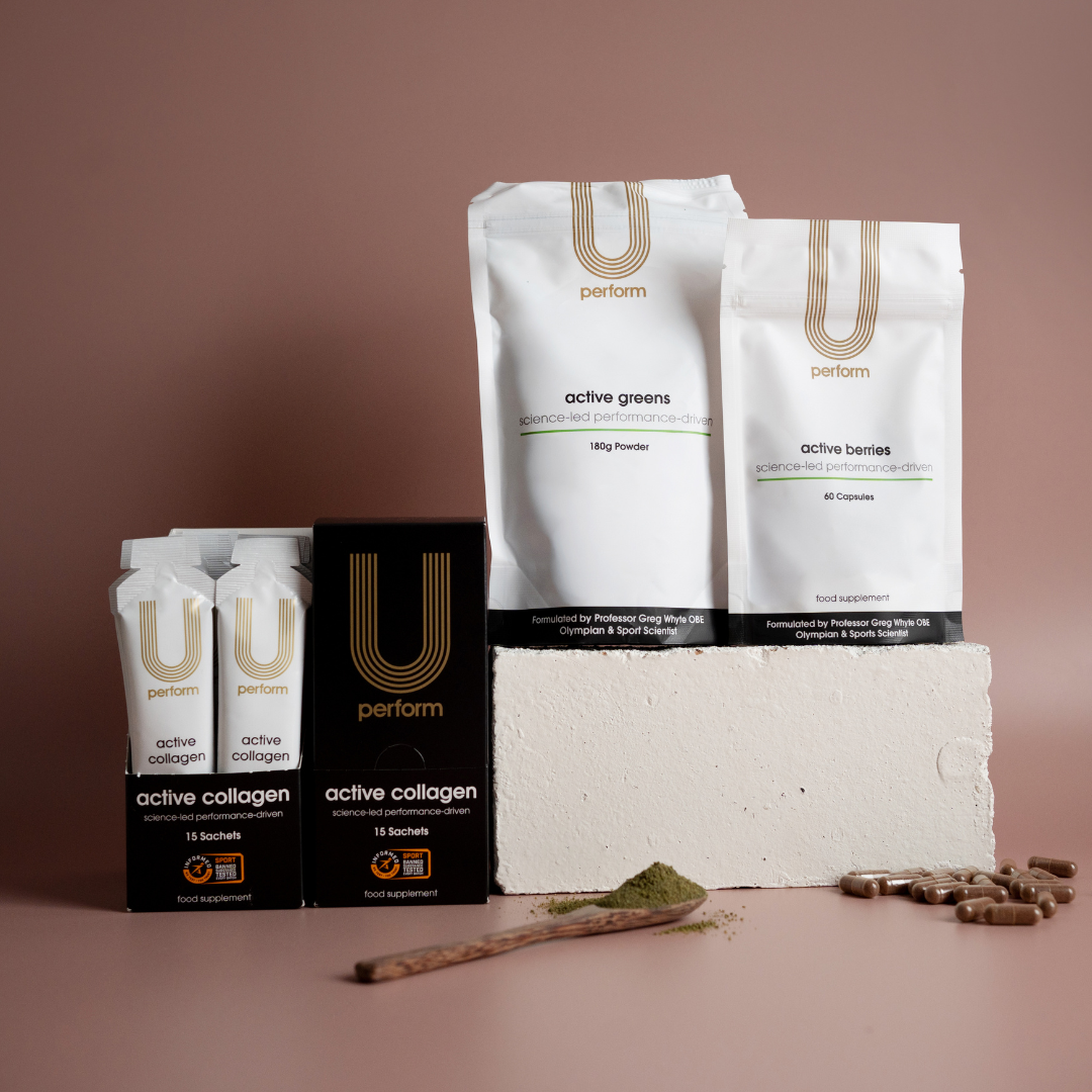 U Perform supplement range displayed against a pink background with a white block, featuring two black boxes and sachets of "active collagen," a white pouch of "active greens" powder, and a smaller pouch of "active berries" capsules. Green powder is spread beside a wooden spoon, and brown capsules are scattered nearby, emphasizing the product forms.