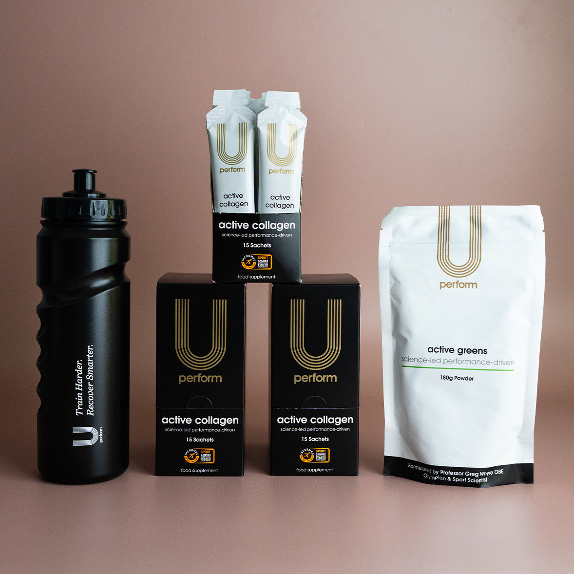 Lineup of U Perform branded products arranged against a muted pink background, including a black sports water bottle, three boxes of active collagen sachets, two sachets displayed on top, and a white pouch of active greens powder. The packaging emphasizes performance, science-led nutrition, and sleek, minimalist branding.